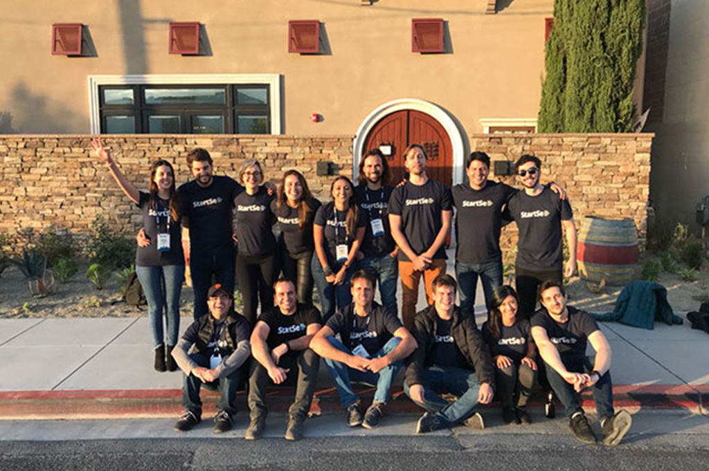 Parte da equipe da StartSe (9 pessoas de pé e 6 pessoas sentadas, numa calçada), todas olhando para a foto, em frente a um lugar com muro baixo feito de tijolos, em San Carlos, Califórnia.