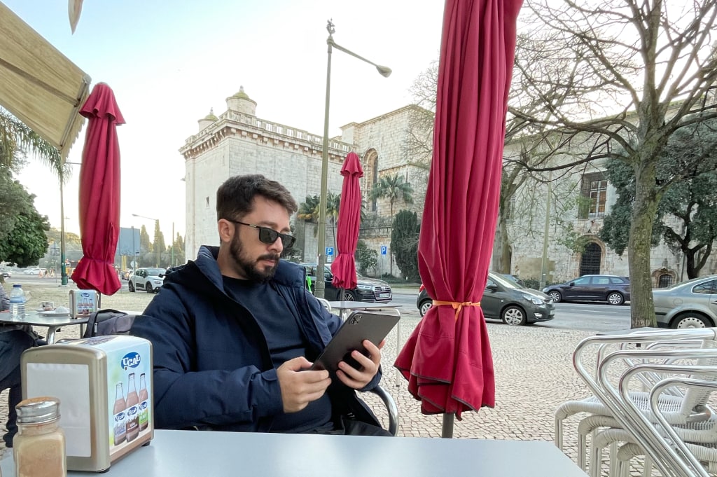 Eder Christian em Lisboa, Portugal. Está sentado numa cadeira, lendo algo no iPad. Ao fundo, uma rua com carros estacionados; céu bem claro.