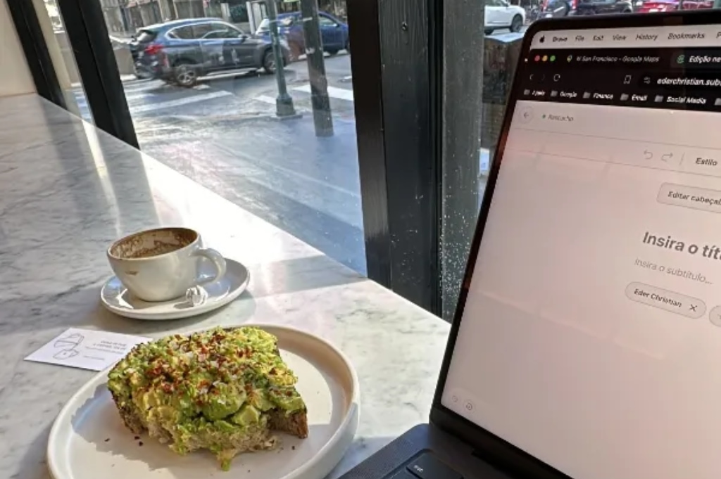 Tela do MacBook, pão com ovo e avocado, e uma xícara de capuccino. Tudo sobre uma mesa branca. Ao fundo dá para ver um pouco de uma rua de San Francisco.