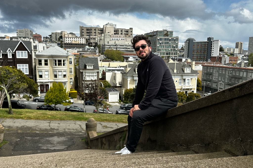 Eder Christian sentado numa escada de cimento com vista de San Francisco ao fundo